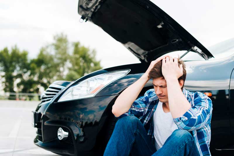 Autoankauf vor Ort – Vertrauen Sie unserem regionalen Service Ein Mann sitzt auf dem Boden neben einem schwarzen Auto mit angehobener Motorhaube und zeigt Anzeichen von Stress und Frustration.